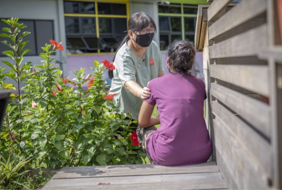 A staff from The Salvation Army wearing a mask supporting a lady sitting on a bench next to a garden